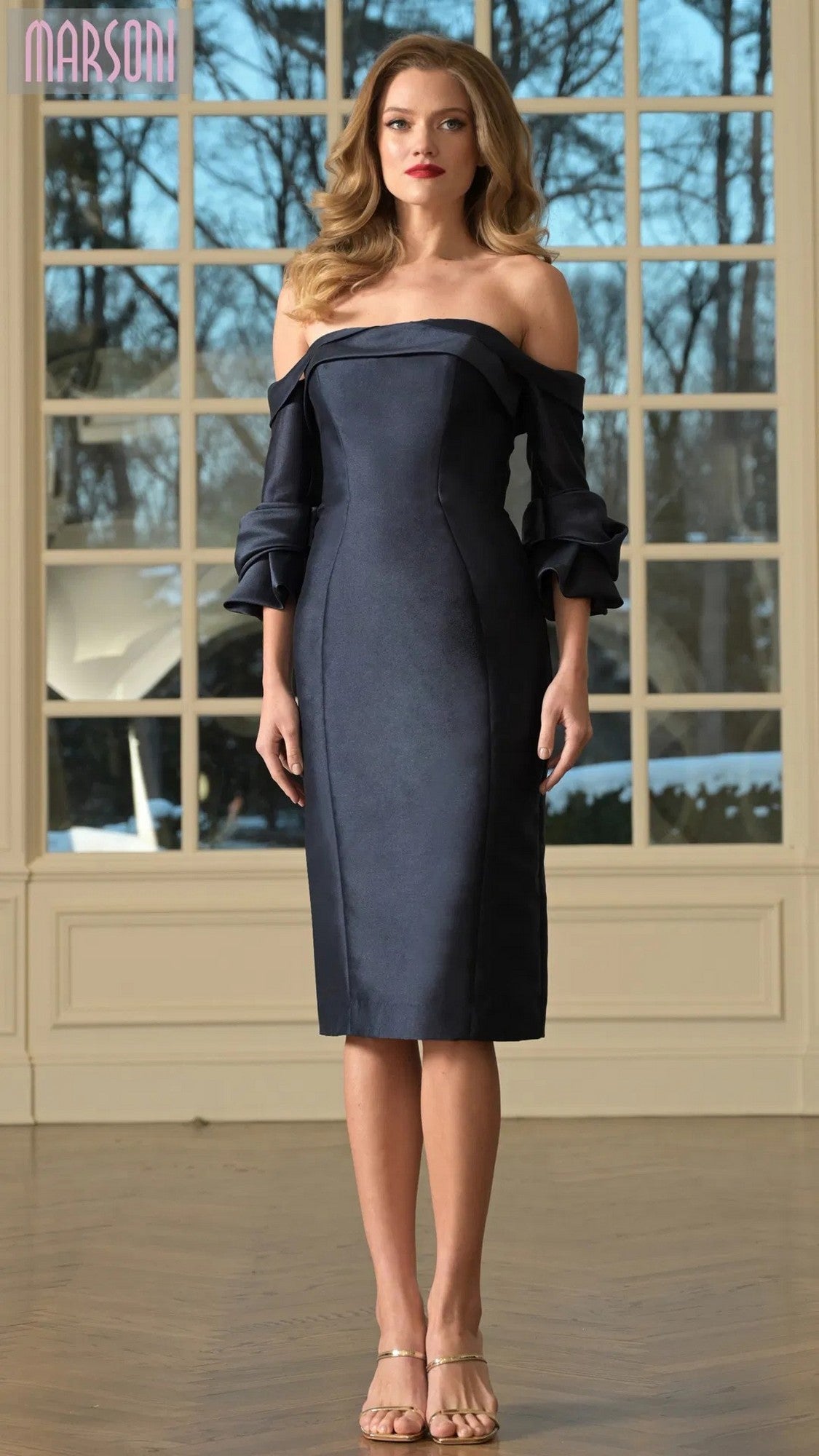 A woman stands indoors wearing the Marsoni MV1370 Knee Length Semi-Formal Dress in navy with ruffled off-the-shoulder sleeves and nude heels. Large windows behind her reveal a view of trees.